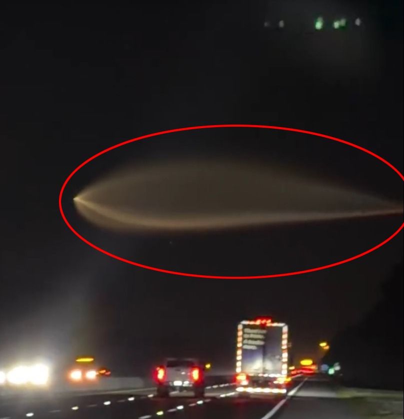 A luminous, elongated object in the night sky above a freeway, seen from a car.
