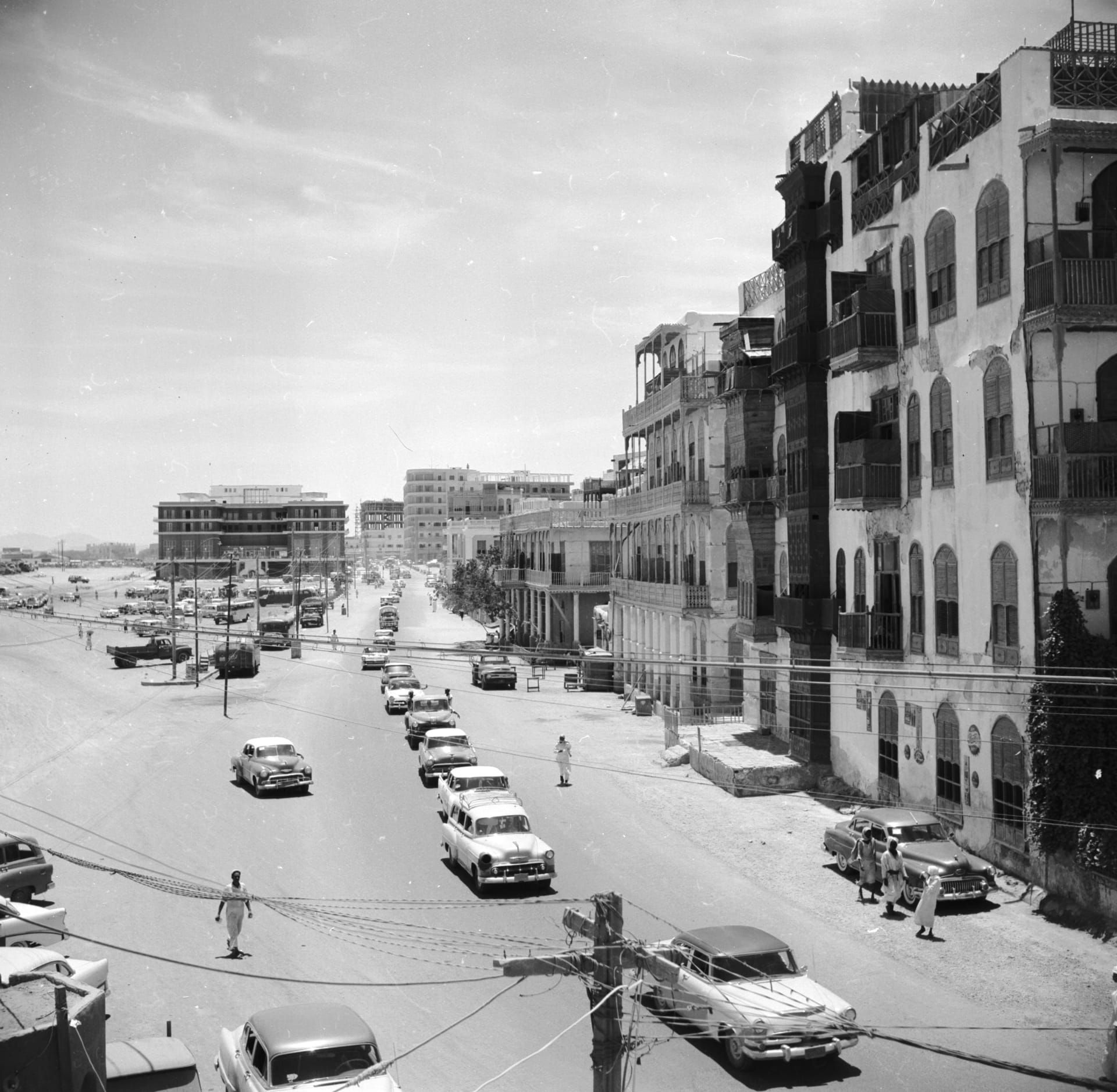 حركة المرور في شوارع جدة، من حوالي ١٩٥٦م. تظهر المباني القديمة بروشانها الخشبية إلى اليمين، بينما ترتفع في الخلفية المباني الإسمنتية الحديثة